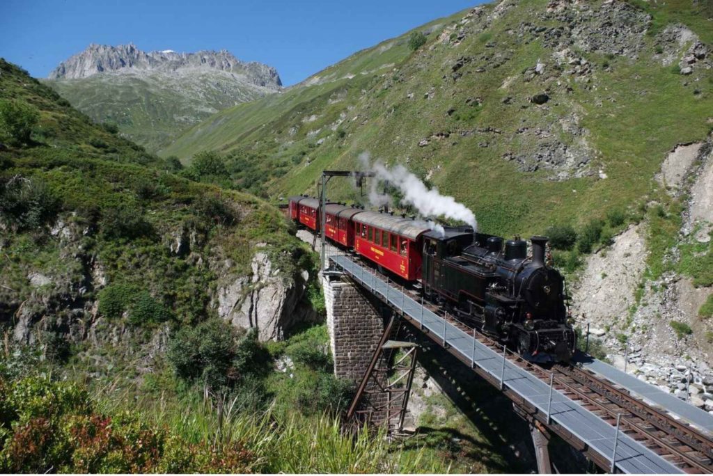 Furka Cogwheel Railway heritage steam locomotive crossing mountain viaduct in Alpine valley landscape, Switzerland