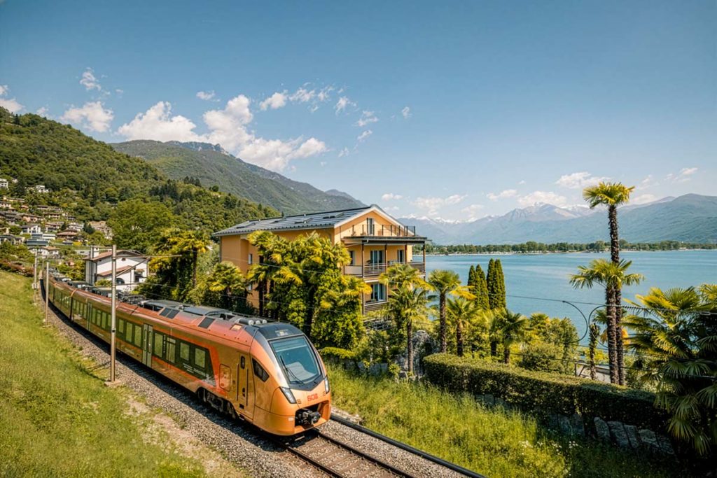 Orange Südostbahn Treno Gottardo train travelling beside Lake Maggiore with palm trees and mountains